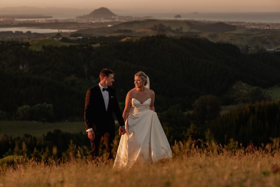 sunset photo of bride and groom walking in field above Tauranga with the ocean views