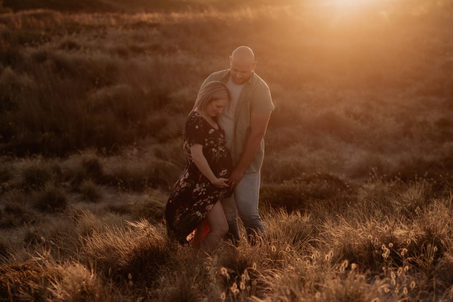 Sun lit lifestyle maternity photos of Mum and Dad looking down at baby