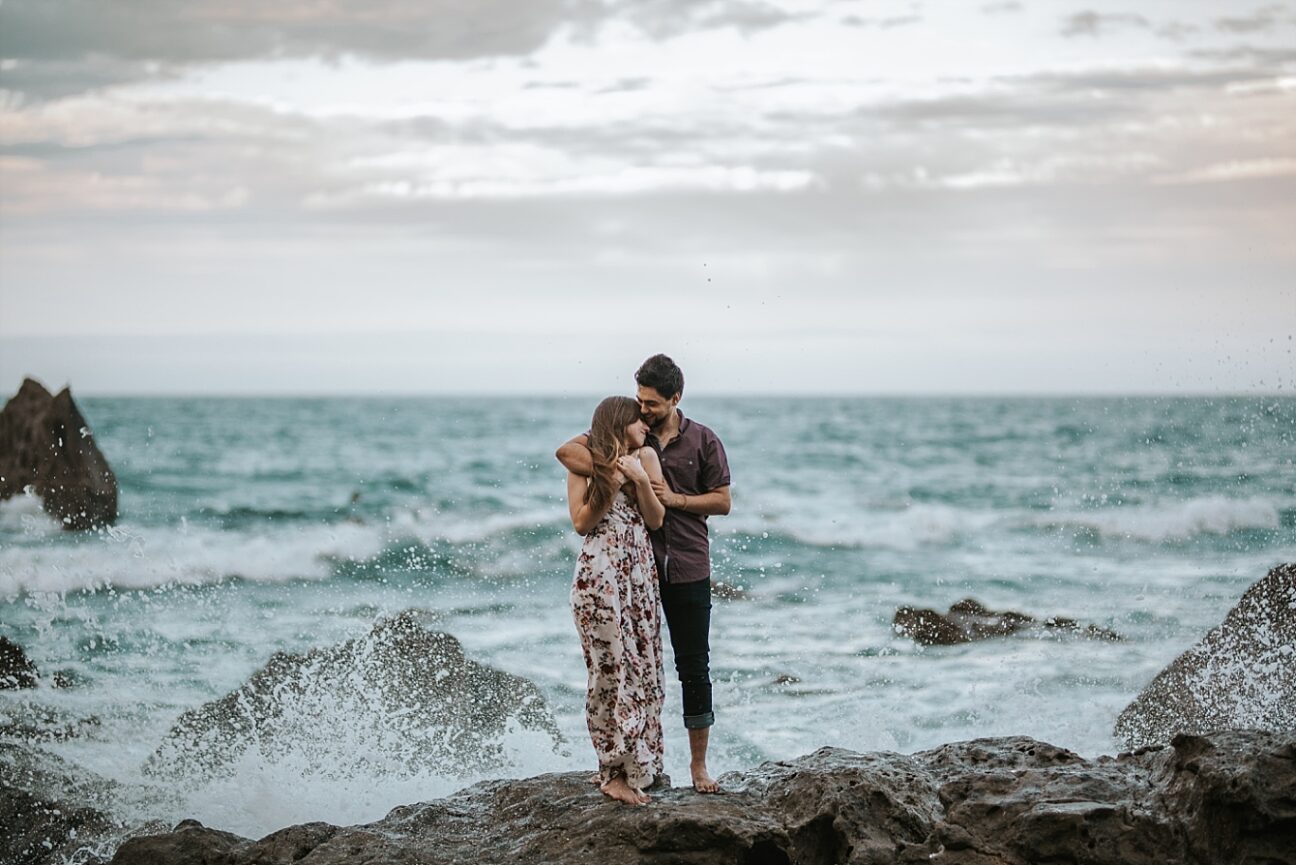 engaged couple cuddle on rocks in front of ocean