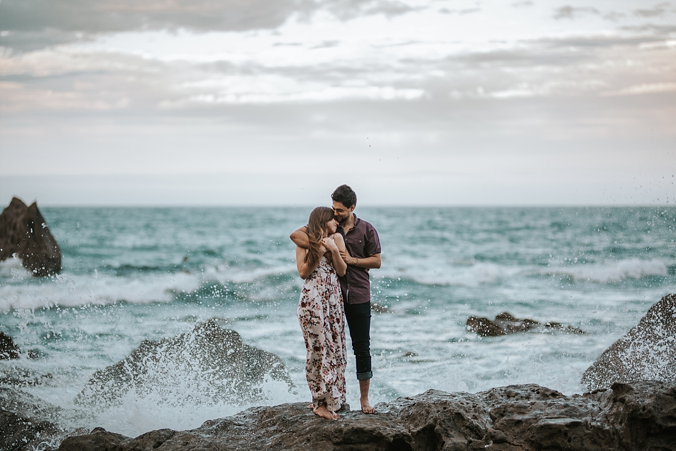 engaged couple cuddle on rocks in front of ocean