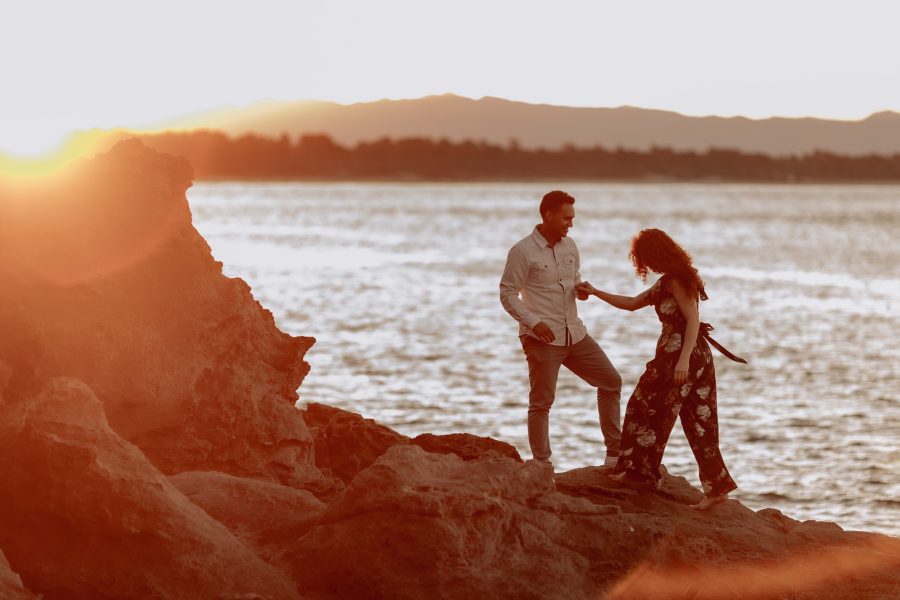 Husband helps wife climb over rocks at golden hour at Tauranga