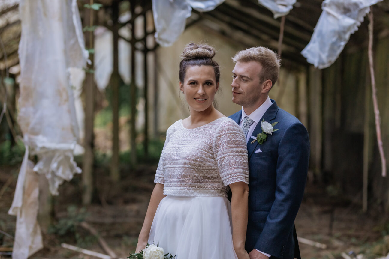 Bride looking at camera with her husband in unused rustic glass house