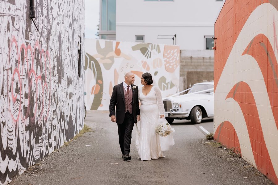 Bridal couple walking hand in hand in front of artwork in back alleys of Mount Maungaui urban wedding