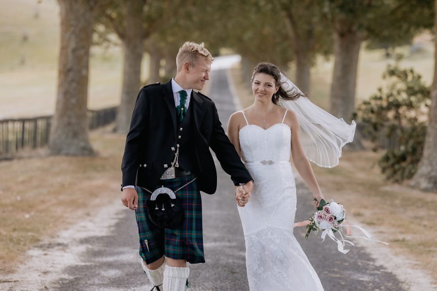 Groom in kilt looks at bride while walking on redbarns waikato driveway