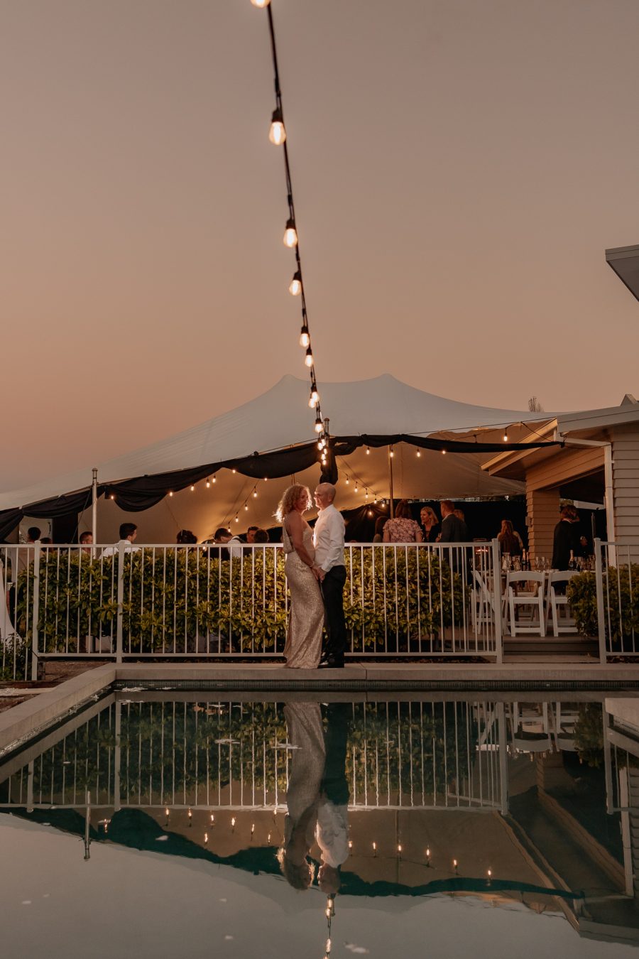 wedding photo by pool at sunset with marquee and celebration lights