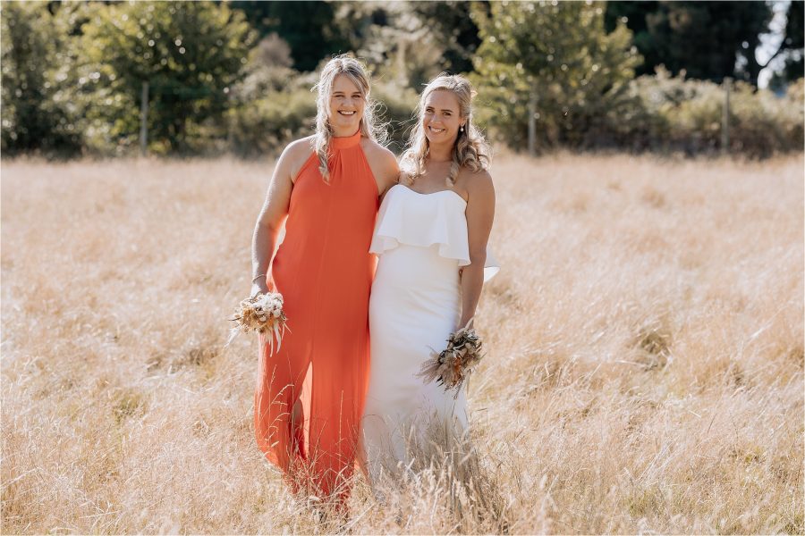 Bride and bridesmaid laughing walking in field, orange dress, summertime