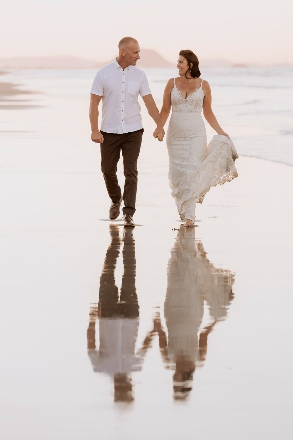 Happy bride and groom casually walking on beach with Mount Maunganui scene