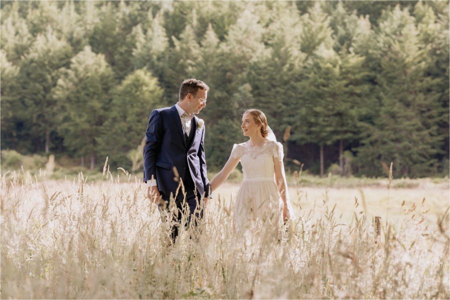 Wedding photo of bride and groom walking through a field of high grass at Old Forest School with trees behind