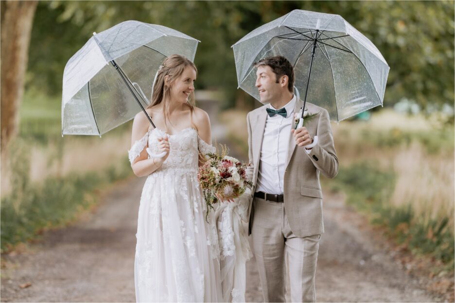Bride and groom holding umbrellas in the rain at Longfords estate driveway