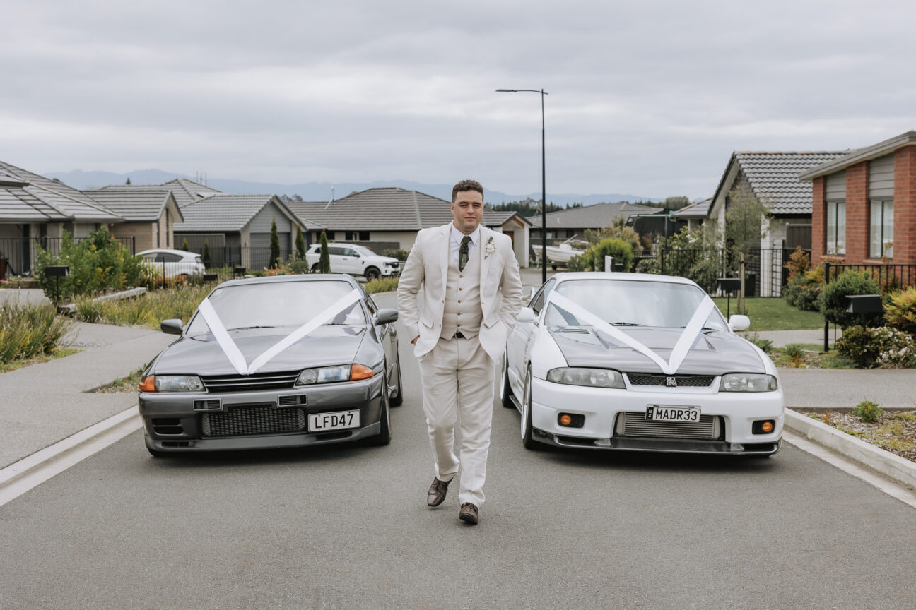 groom walking on road in front of wedding cars