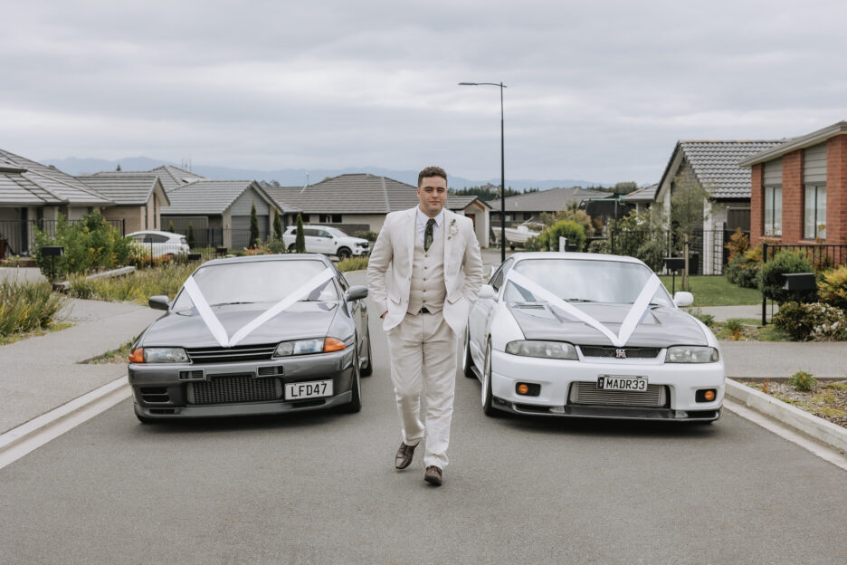 groom walking on road in front of wedding cars