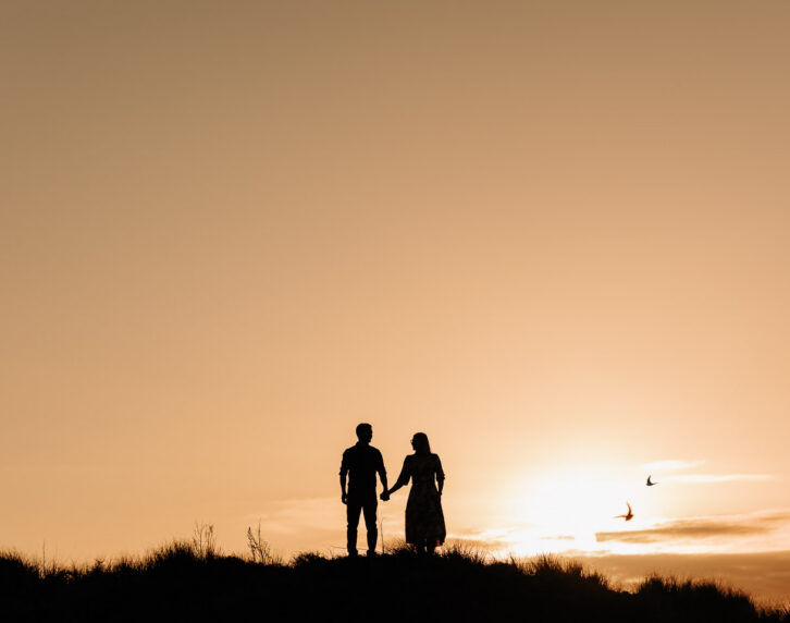 engaged couple standing at sunset Papamoa sand dunes with birds flying