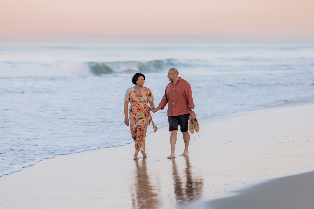 Lady in floral dress with man in red shirt walking beach at sunset during photo session