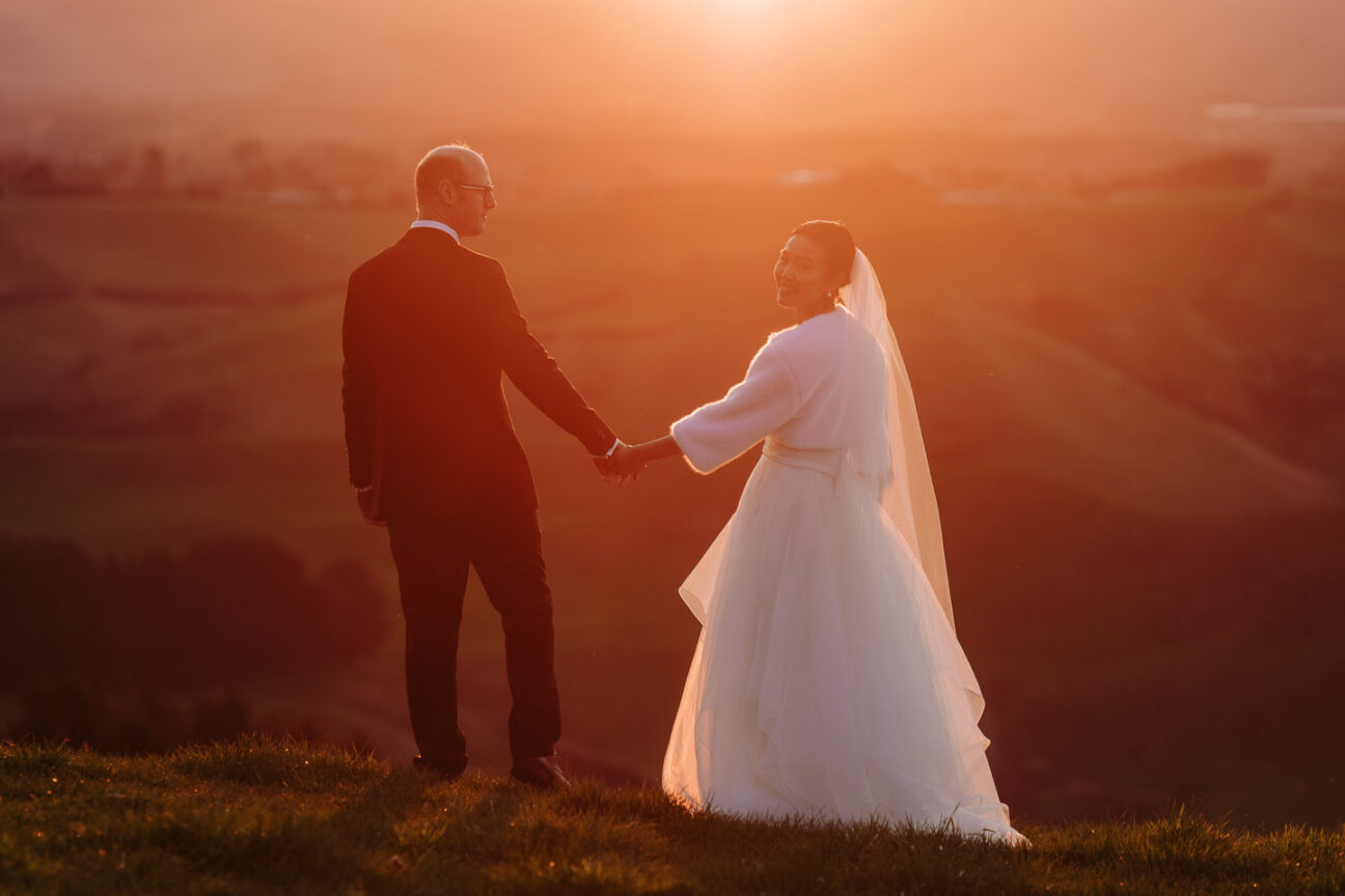 Bride looks over shoulder walking over hill as sun sets over Tauranga hills