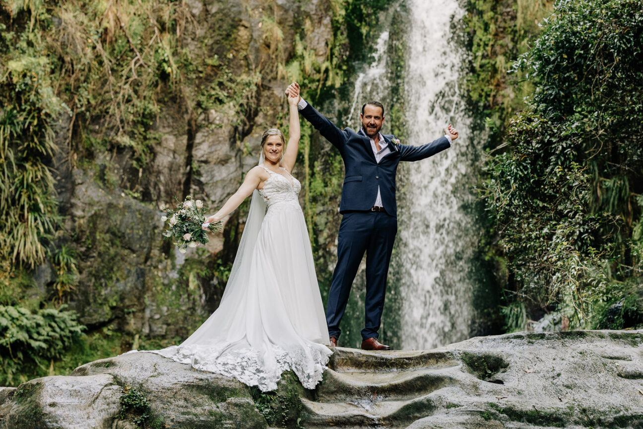 elopement couple just married in front of waterfall hold up their arms in celebration