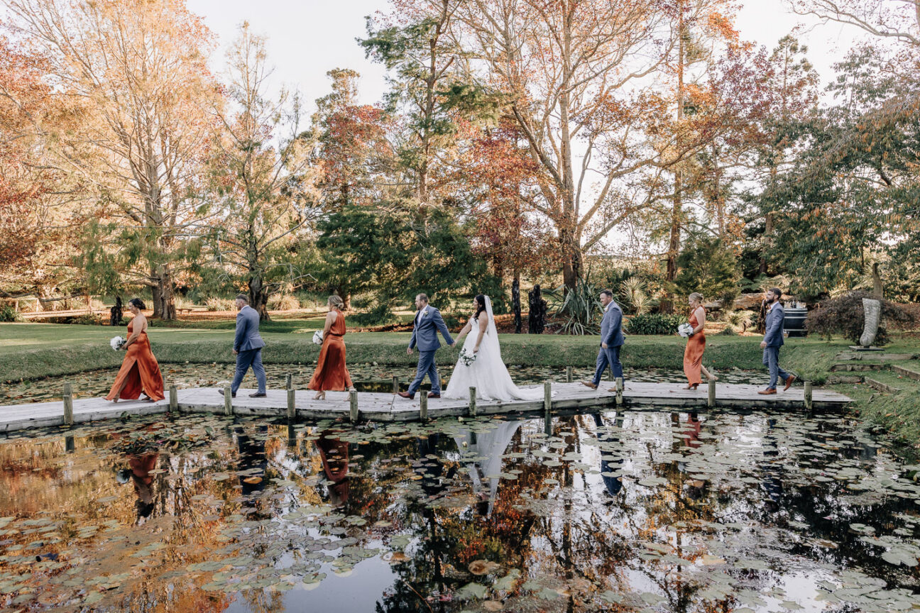 wedding party walk across pond board walk at waterlily gardens Waihi in autumn orange dresses
